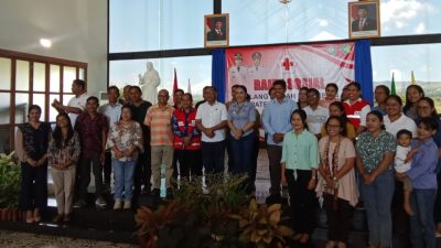 Bupati SBD, Rektor Unika Weetabula, dan tim PMI, berdiri di panggung Aula Maria Ratu Damai. Di latar belakang terdapat patung Yesus Kristus, spanduk merah Bakti Sosial PMI, dan foto-foto resmi pejabat negara. Suasana hangat dan ramah.