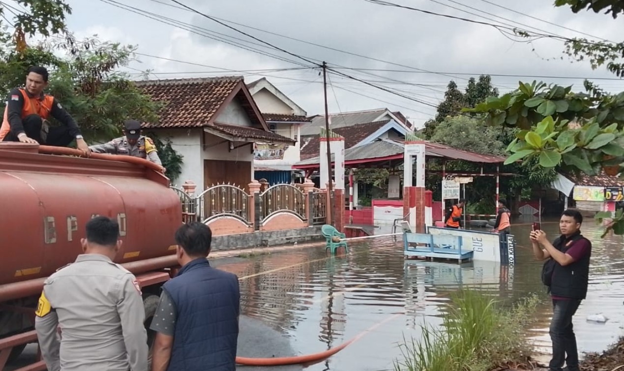 Foto tingkat mata dari jalan raya yang tergenang banjir di Sukaraya, Baturaja, menampakkan truk tangki merah BPBD, petugas polisi, dan warga di depan rumah-rumah penduduk.