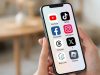 Sebuah foto close-up tangan seseorang yang memegang iPhone modern. Layar ponsel menampilkan grid ikon aplikasi untuk YouTube, TikTok, Facebook, Instagram, Threads, X, Bigo Live, dan Roblox yang tertata rapi. Ikon-ikon tersebut berwarna cerah di atas latar belakang buram interior rumah yang bersahaja.