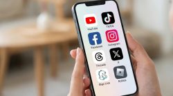 Sebuah foto close-up tangan seseorang yang memegang iPhone modern. Layar ponsel menampilkan grid ikon aplikasi untuk YouTube, TikTok, Facebook, Instagram, Threads, X, Bigo Live, dan Roblox yang tertata rapi. Ikon-ikon tersebut berwarna cerah di atas latar belakang buram interior rumah yang bersahaja.