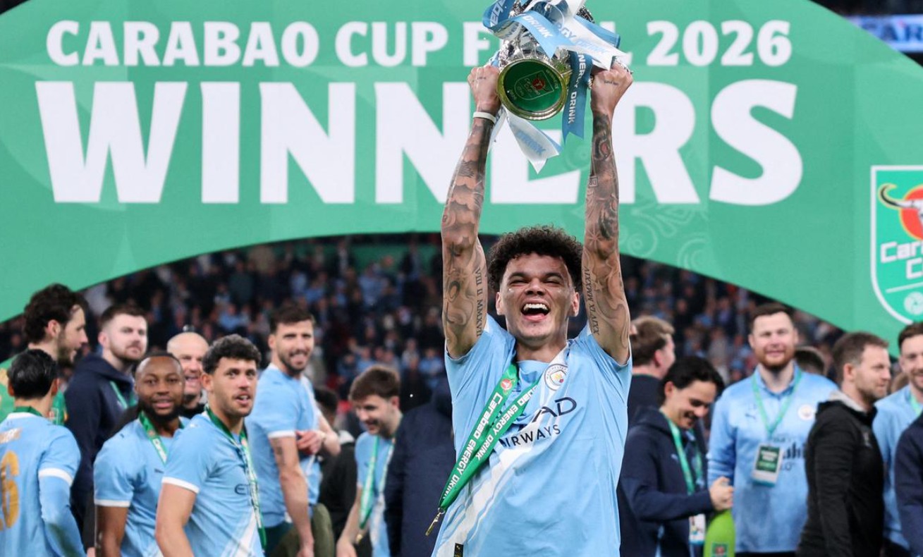 pemain muda Manchester City dengan banyak tato di lengan, Nico O'Reilly, merayakan kemenangan sendirian di depan spanduk panggung bertuliskan "CARABAO CUP FINAL 2026 WINNERS".