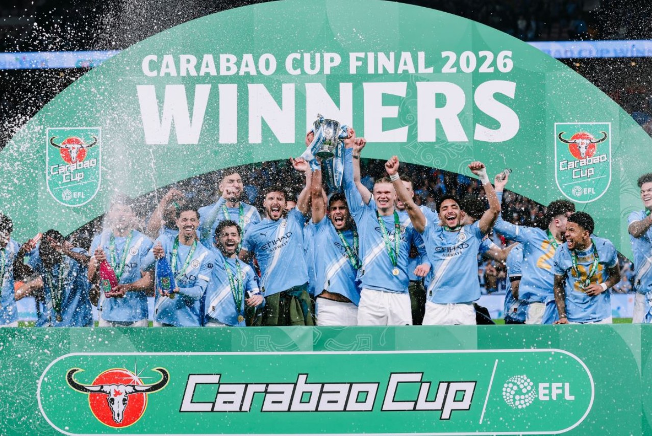 pemain Manchester City berkumpul di atas panggung merayakan kemenangan. Spanduk besar di belakang mereka bertuliskan "CARABAO CUP FINAL 2026 WINNERS". Beberapa pemain mengangkat trofi bersama-sama, dan seorang pemain menyemprotkan sampanye ke udara di tengah kerumunan yang gembira. Logo Carabao Cup EFL terlihat di pagar depan.