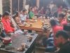 Pemandangan dalam ruangan di malam hari yang hangat, memperlihatkan sekelompok besar anak muda duduk mengelilingi meja-meja kayu untuk rapat sosial atau diskusi. Meja-meja itu penuh dengan cangkir-cangkir kecil, dan ada kue ulang tahun berbentuk tabung berwarna merah di meja terpisah di latar depan. Beberapa orang mengenakan kemeja merah yang serasi di sisi kanan.