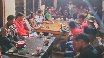 Pemandangan dalam ruangan di malam hari yang hangat, memperlihatkan sekelompok besar anak muda duduk mengelilingi meja-meja kayu untuk rapat sosial atau diskusi. Meja-meja itu penuh dengan cangkir-cangkir kecil, dan ada kue ulang tahun berbentuk tabung berwarna merah di meja terpisah di latar depan. Beberapa orang mengenakan kemeja merah yang serasi di sisi kanan.