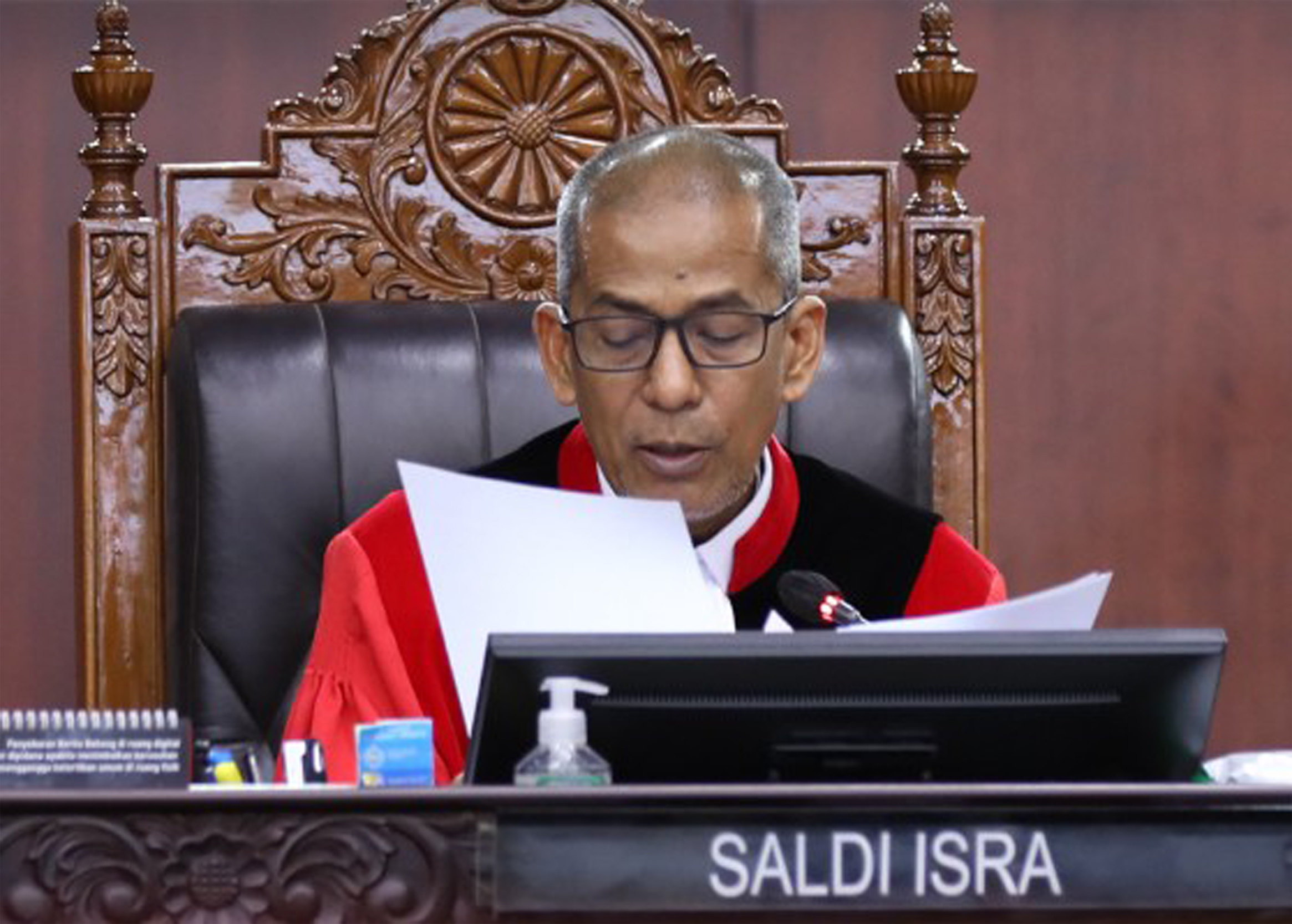 Hakim Mahkamah Konstitusi Saldi Isra mengenakan toga hitam dan merah sedang fokus membaca dokumen putusan di persidangan.