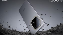 Tampilan render produk smartphone iQOO 15 mini berwarna abu-abu metallic dengan modul kamera bulat menonjol. Perangkat ini bersandar di atas permukaan batu kasar yang retak, dengan serpihan batu kecil melayang secara dinamis di sekelilingnya.