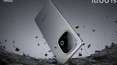 Tampilan render produk smartphone iQOO 15 mini berwarna abu-abu metallic dengan modul kamera bulat menonjol. Perangkat ini bersandar di atas permukaan batu kasar yang retak, dengan serpihan batu kecil melayang secara dinamis di sekelilingnya.