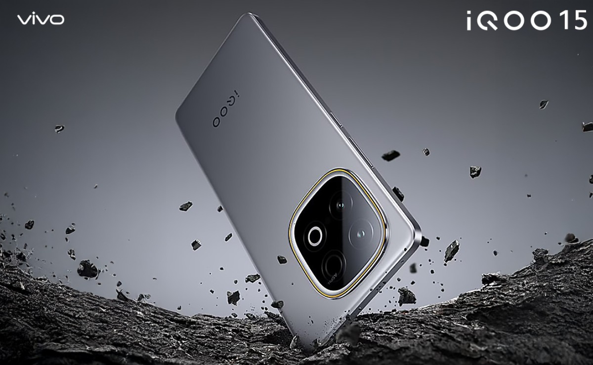 Tampilan render produk smartphone iQOO 15 berwarna abu-abu metallic dengan modul kamera bulat menonjol. Perangkat ini bersandar di atas permukaan batu kasar yang retak, dengan serpihan batu kecil melayang secara dinamis di sekelilingnya.