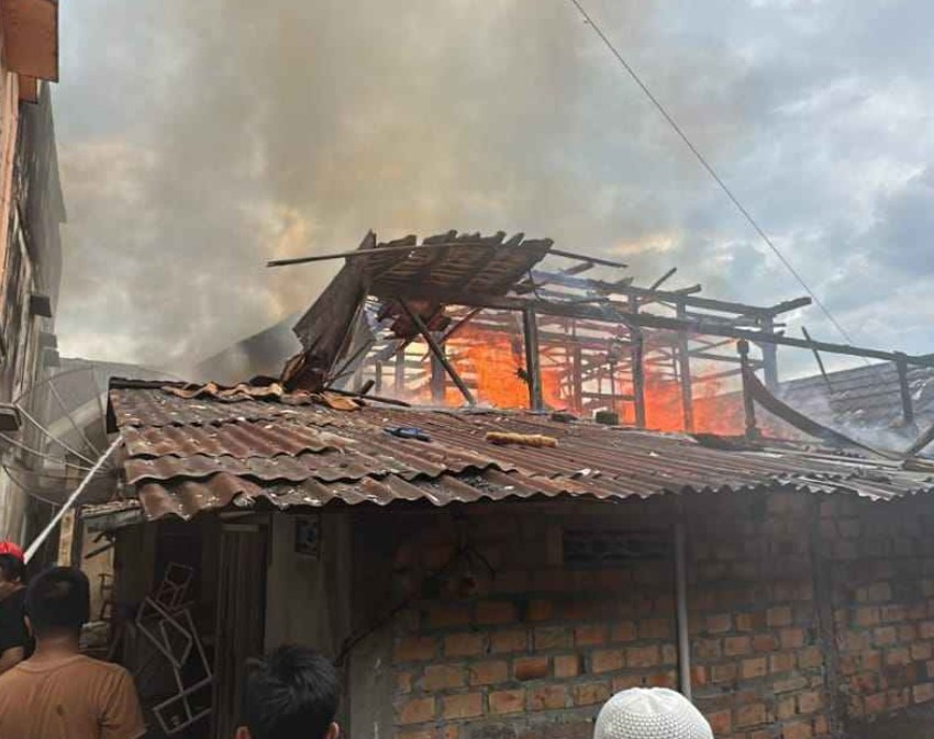 Rumah bata dengan atap asbes hancur sebagian dan api terlihat membakar rangka kayu bagian dalamnya. Asap tebal mengepul dari atas bangunan yang rusak, disaksikan oleh warga dari kejauhan.