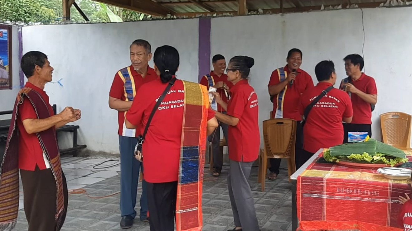 ekelompok anggota Punguan Batak Muaradua, OKU Selatan, berinteraksi dan tertawa riang dalam perayaan HUT ke-32. Mereka mengenakan kemeja merah seragam berlogo dan kain adat ulos di bahu, dengan meja perjamuan tumpeng dan penanda acara di latar belakang.