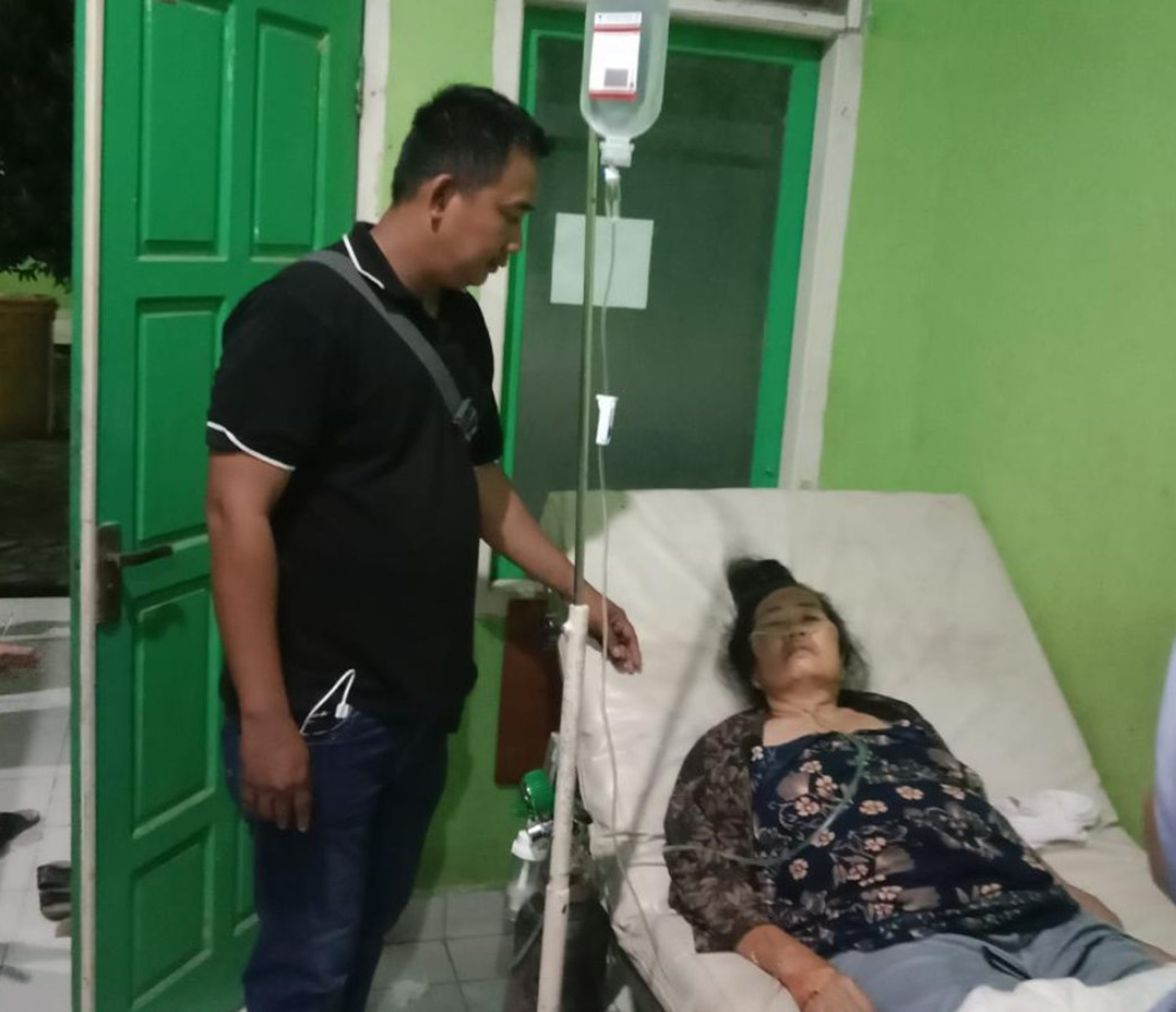 Seorang wanita paruh baya berbaring di tempat tidur rumah sakit dengan kepala bersandar pada bantal tinggi. Ia mengenakan kanula oksigen hidung dan infus IV terpasang.