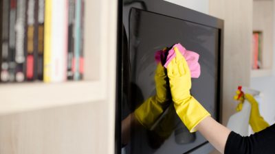 Seseorang memakai sarung tangan karet kuning sedang mengelap layar TV datar menggunakan kain merah muda sambil memegang botol semprotan.