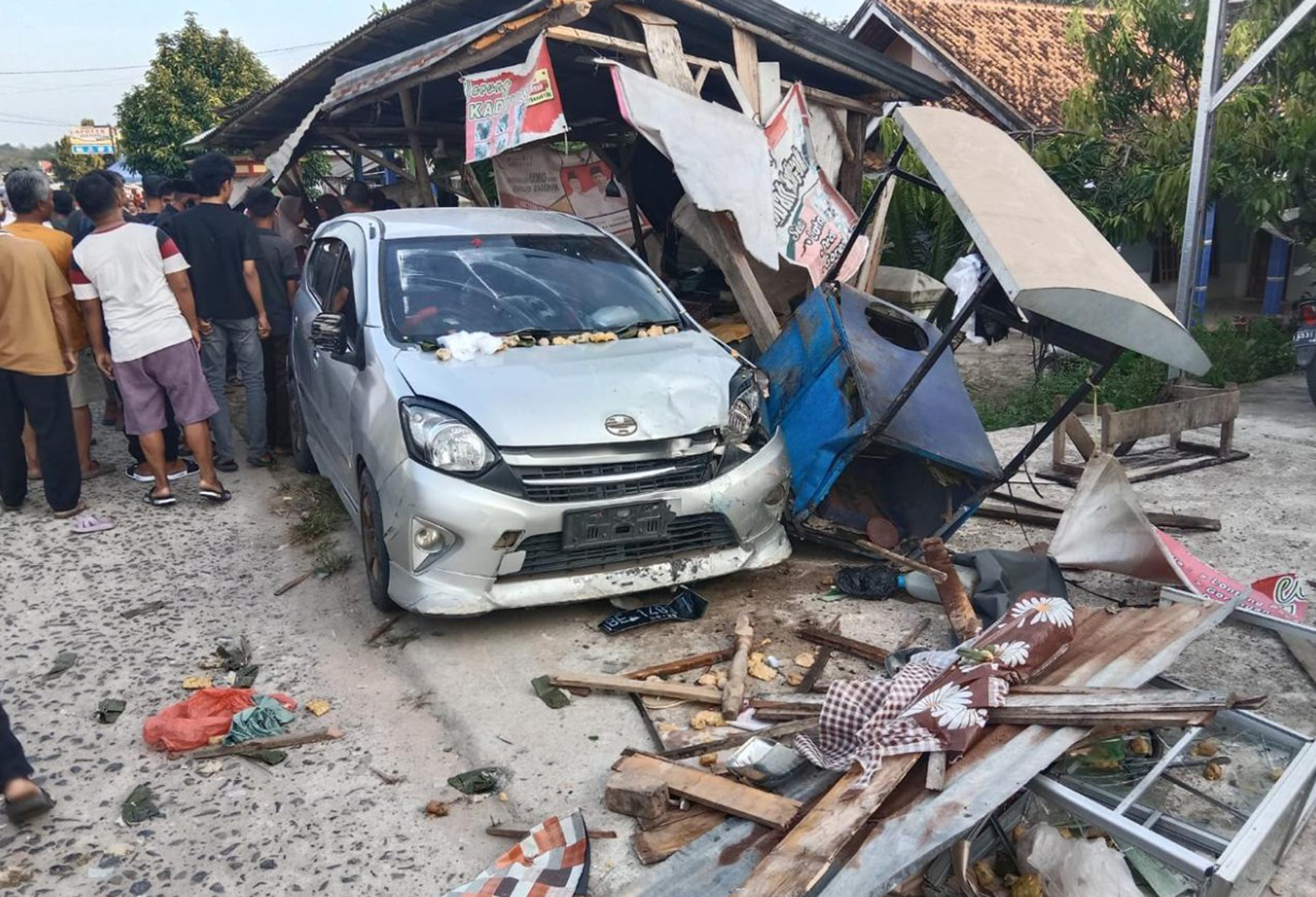 Tampilan dekat bagian depan mobil Toyota Agya perak yang rusak parah, tertanam di dalam puing-puing warung kayu dan atap seng yang hancur. Kayu patah, gerobak biru yang terbalik, dan barang-barang berserakan di tanah. Sekelompok orang berkumpul di sebelah kiri, mengamati pemandangan tersebut.