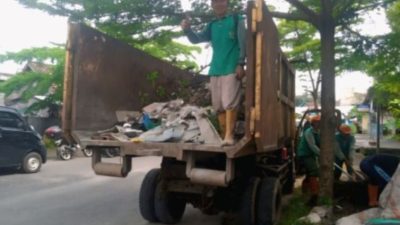 petugas kebersihan DLH dengan sepatu bot oranye bekerja keras membersihkan tumpukan sampah di area terbuka. Dua pengawas berdiri di latar belakang di dekat pagar dan bangunan.