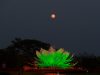 Sebuah patung teratai raksasa yang menyala hijau cerah di malam hari, diposisikan di tengah taman dengan bulan purnama berwarna merah muda/tembaga di langit gelap di atasnya. Siluet pohon-pohon gelap di latar belakang.