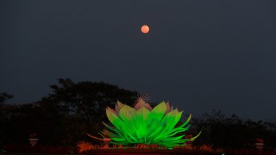Sebuah patung teratai raksasa yang menyala hijau cerah di malam hari, diposisikan di tengah taman dengan bulan purnama berwarna merah muda/tembaga di langit gelap di atasnya. Siluet pohon-pohon gelap di latar belakang.