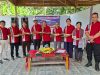 Sekelompok anggota Punguan Batak Muaradua, OKU Selatan, berinteraksi dan tertawa riang dalam perayaan HUT ke-32. Mereka mengenakan kemeja merah seragam berlogo dan kain adat ulos di bahu, dengan meja perjamuan tumpeng dan penanda acara di latar belakang.