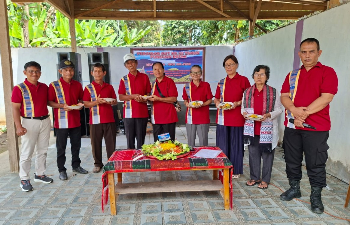 Sekelompok anggota Punguan Batak Muaradua, OKU Selatan, berinteraksi dan tertawa riang dalam perayaan HUT ke-32. Mereka mengenakan kemeja merah seragam berlogo dan kain adat ulos di bahu, dengan meja perjamuan tumpeng dan penanda acara di latar belakang.