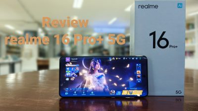 Kotak produk secara menonjol menampilkan teks 'realme' dan '16 Pro+ 5G' serta logo 'Ai' kecil. Terdapat teks overlay berwarna tembaga di sisi kiri bertuliskan 'Review realme 16 Pro+ 5G'. Latar belakang adalah lingkungan kantor yang terang dan buram.