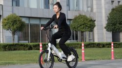 Seorang wanita mengendarai sepeda listrik lipat berwarna putih di jalan raya perkotaan.