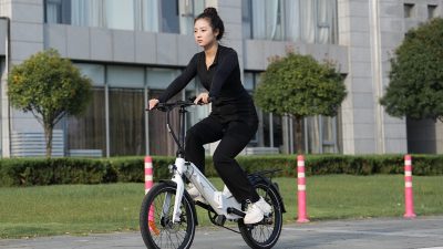 Seorang wanita mengendarai sepeda listrik lipat berwarna putih di jalan raya perkotaan.