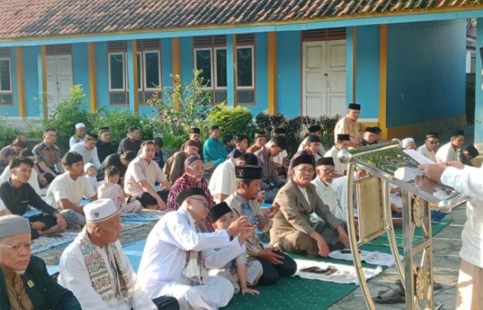 Sebuah foto luar ruangan yang memperlihatkan ratusan jemaah pria Muslim duduk tertib di atas sajadah untuk menjalankan shalat berjamaah di halaman depan sebuah madrasah bercat biru cerah dengan aksen kuning.