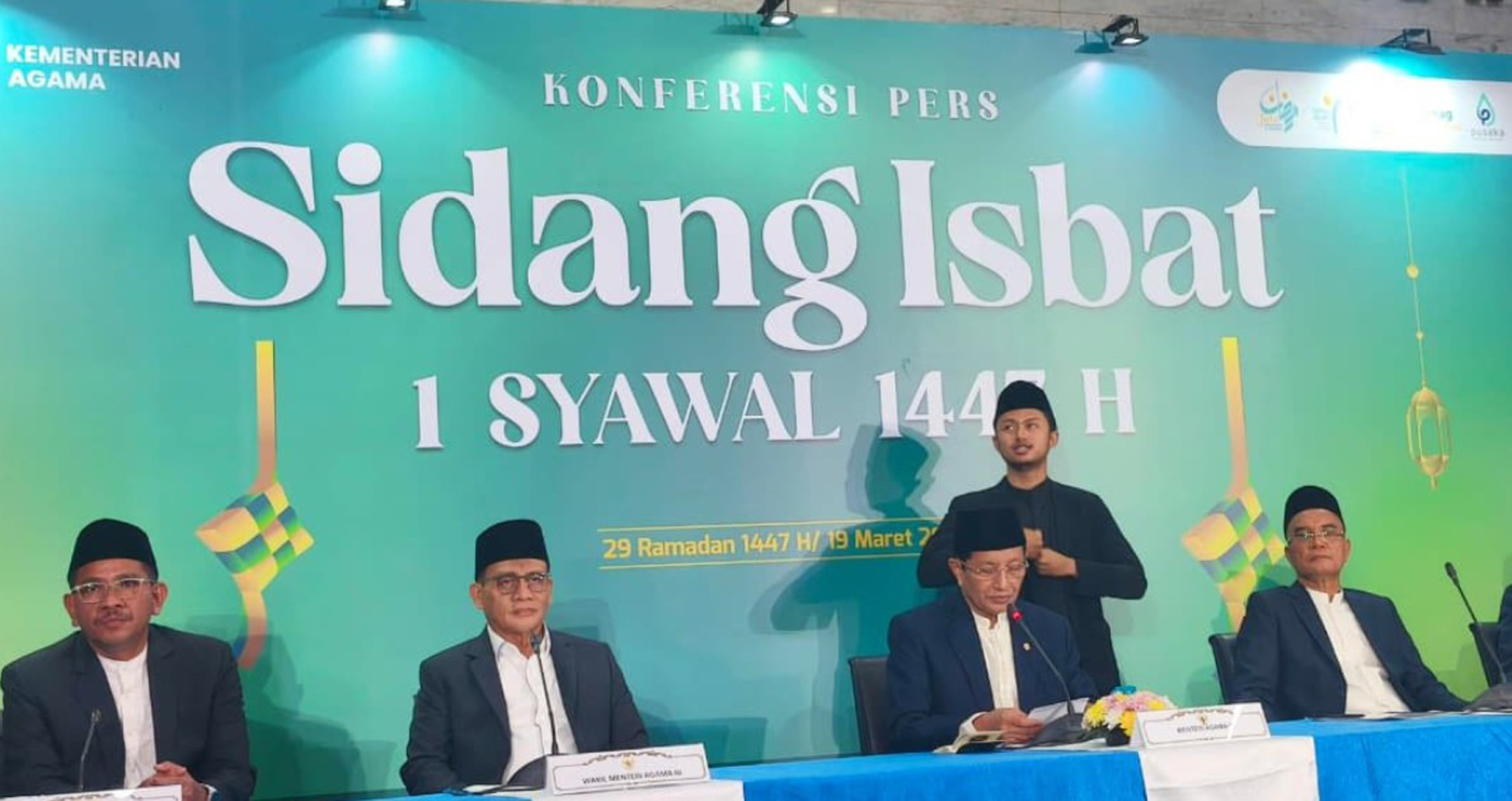 Sekelompok pria berpeci hitam sedang duduk di sebuah meja panjang dengan latar belakang hijau bertuliskan "Sidang Isbat 1 Syawal 1447 H". Salah satu pria berdiri dan berjarak dengan pria yang lainnya, sementara sisanya duduk. Terdapat hiasan ketupat di sebelah kiri dan kanan, serta logo "KEMENTERIAN AGAMA" di bagian atas.