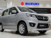 Sebuah foto tiga perempat depan dari mobil kompak Suzuki Wagon R perak yang diparkir di lantai ubin putih di dalam dealer mobil Suzuki yang bersih dan modern, dengan beberapa logo Suzuki merah dan biru terlihat di dinding latar belakang.