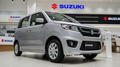 Sebuah foto tiga perempat depan dari mobil kompak Suzuki Wagon R perak yang diparkir di lantai ubin putih di dalam dealer mobil Suzuki yang bersih dan modern, dengan beberapa logo Suzuki merah dan biru terlihat di dinding latar belakang.