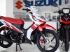 Di dalam showroom Suzuki yang terang dan bersih, dua sepeda motor underbone Suzuki Smash Fi dipajang. Motor di sebelah kiri berwarna putih dengan grafis merah dan hitam, dilengkapi kursi merah cerah, shock absorber belakang merah, dan kaliper rem depan merah.
