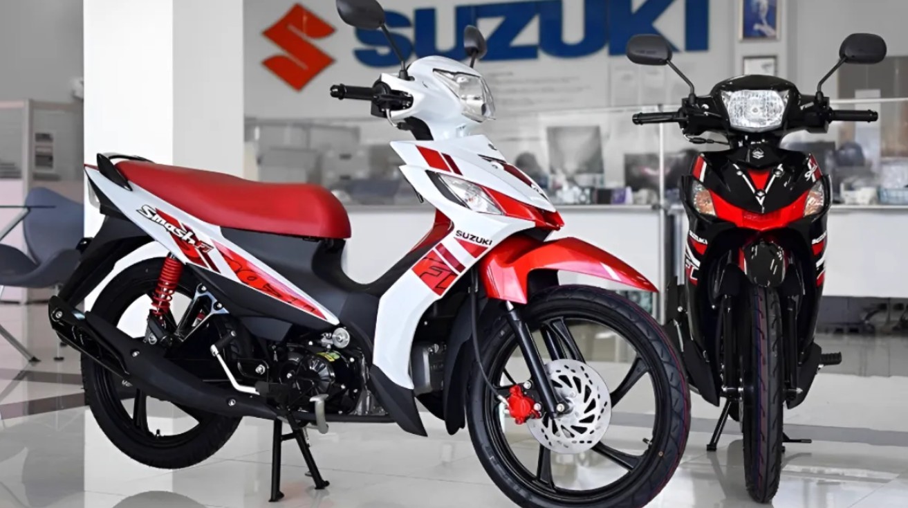 Di dalam showroom Suzuki yang terang dan bersih, dua sepeda motor underbone Suzuki Smash Fi dipajang. Motor di sebelah kiri berwarna putih dengan grafis merah dan hitam, dilengkapi kursi merah cerah, shock absorber belakang merah, dan kaliper rem depan merah.