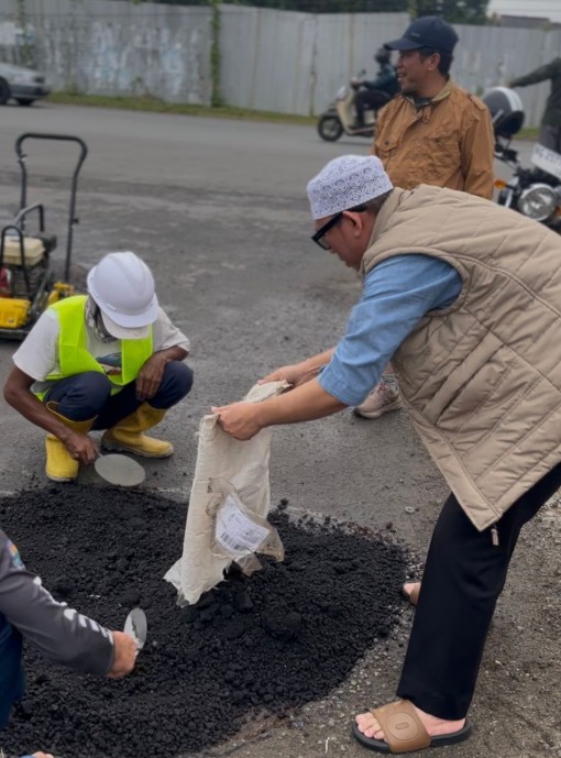 "Wujud komitmen pelayanan publik. Sambil memantau, Bupati OKU Teddy Meilwansyah berinteraksi dengan warga yang melintas (pria berjaket cokelat di belakang) saat proses penambalan jalan rusak. Perbaikan ini diprioritaskan pada akses vital sehari-hari."