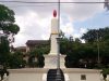 Sebuah monumen besar berbentuk lilin putih dengan nyala api merah di puncaknya dan pita emas, berdiri di atas dasar beton bertingkat dengan tangga kecil. Di depannya berdiri pilar lampu jalan logam dekoratif berwarna gelap. Di latar belakang, terdapat pepohonan hijau lebat dan sebuah bangunan kolonial tua di bawah langit berawan. Kabel listrik melintang di bagian atas bingkai.