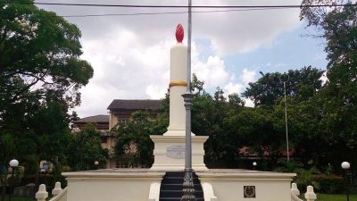 Sebuah monumen besar berbentuk lilin putih dengan nyala api merah di puncaknya dan pita emas, berdiri di atas dasar beton bertingkat dengan tangga kecil. Di depannya berdiri pilar lampu jalan logam dekoratif berwarna gelap. Di latar belakang, terdapat pepohonan hijau lebat dan sebuah bangunan kolonial tua di bawah langit berawan. Kabel listrik melintang di bagian atas bingkai.