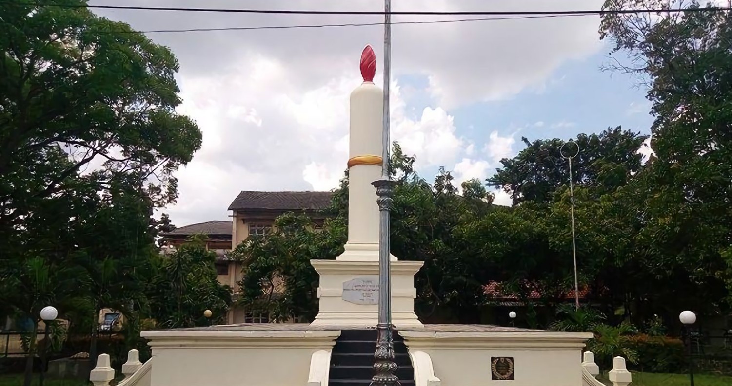 Sebuah monumen besar berbentuk lilin putih dengan nyala api merah di puncaknya dan pita emas, berdiri di atas dasar beton bertingkat dengan tangga kecil. Di depannya berdiri pilar lampu jalan logam dekoratif berwarna gelap. Di latar belakang, terdapat pepohonan hijau lebat dan sebuah bangunan kolonial tua di bawah langit berawan. Kabel listrik melintang di bagian atas bingkai.