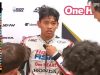 Gambar close-up pembalap muda Indonesia Veda Ega Pratama di area pit tim Honda Team Asia. Seorang asisten sedang menyesuaikan bagian dada setelan balapnya yang berwarna putih. Veda terlihat lelah dan sedikit kecewa, dengan rambut berantakan. Di sisi kiri layar terdapat overlay grafis siaran langsung Moto3 yang menampilkan informasi "LAP 8 OF 14" dan klasemen sementara. Daftar lima besar yang terlihat di grafik adalah MQUI, GPIN, VPER, ACAR, dan AFER. Latar belakang memiliki logo-logo sponsor tim, seperti Honda, HRC, dan One HEART. Logo SPOTV NOW LIVE terlihat di kanan atas.
