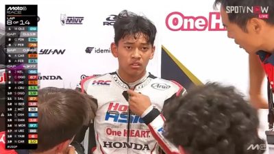 Gambar close-up pembalap muda Indonesia Veda Ega Pratama di area pit tim Honda Team Asia. Seorang asisten sedang menyesuaikan bagian dada setelan balapnya yang berwarna putih. Veda terlihat lelah dan sedikit kecewa, dengan rambut berantakan. Di sisi kiri layar terdapat overlay grafis siaran langsung Moto3 yang menampilkan informasi "LAP 8 OF 14" dan klasemen sementara. Daftar lima besar yang terlihat di grafik adalah MQUI, GPIN, VPER, ACAR, dan AFER. Latar belakang memiliki logo-logo sponsor tim, seperti Honda, HRC, dan One HEART. Logo SPOTV NOW LIVE terlihat di kanan atas.