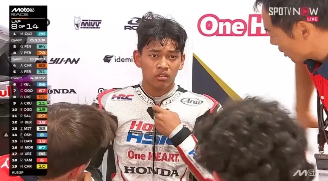 Gambar close-up pembalap muda Indonesia Veda Ega Pratama di area pit tim Honda Team Asia. Seorang asisten sedang menyesuaikan bagian dada setelan balapnya yang berwarna putih. Veda terlihat lelah dan sedikit kecewa, dengan rambut berantakan. Di sisi kiri layar terdapat overlay grafis siaran langsung Moto3 yang menampilkan informasi "LAP 8 OF 14" dan klasemen sementara. Daftar lima besar yang terlihat di grafik adalah MQUI, GPIN, VPER, ACAR, dan AFER. Latar belakang memiliki logo-logo sponsor tim, seperti Honda, HRC, dan One HEART. Logo SPOTV NOW LIVE terlihat di kanan atas.
