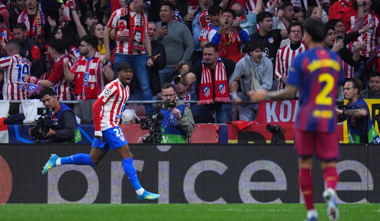 Seorang pemain sepak bola Atletico Madrid (Ademola Lookman, jersey no. 22) berlari dan berteriak merayakan gol di hadapan pendukung yang bersorak. João Cancelo dari Barcelona (jersey no. 2) berdiri di latar depan dengan punggung menghadap kamera, tampak kecewa. Papan iklan LED terlihat.