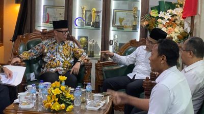 Bupati OKU berbaju batik dan Bupati Bantul berbaju putih duduk berdiskusi di ruang pertemuan dengan latar belakang lemari piala.