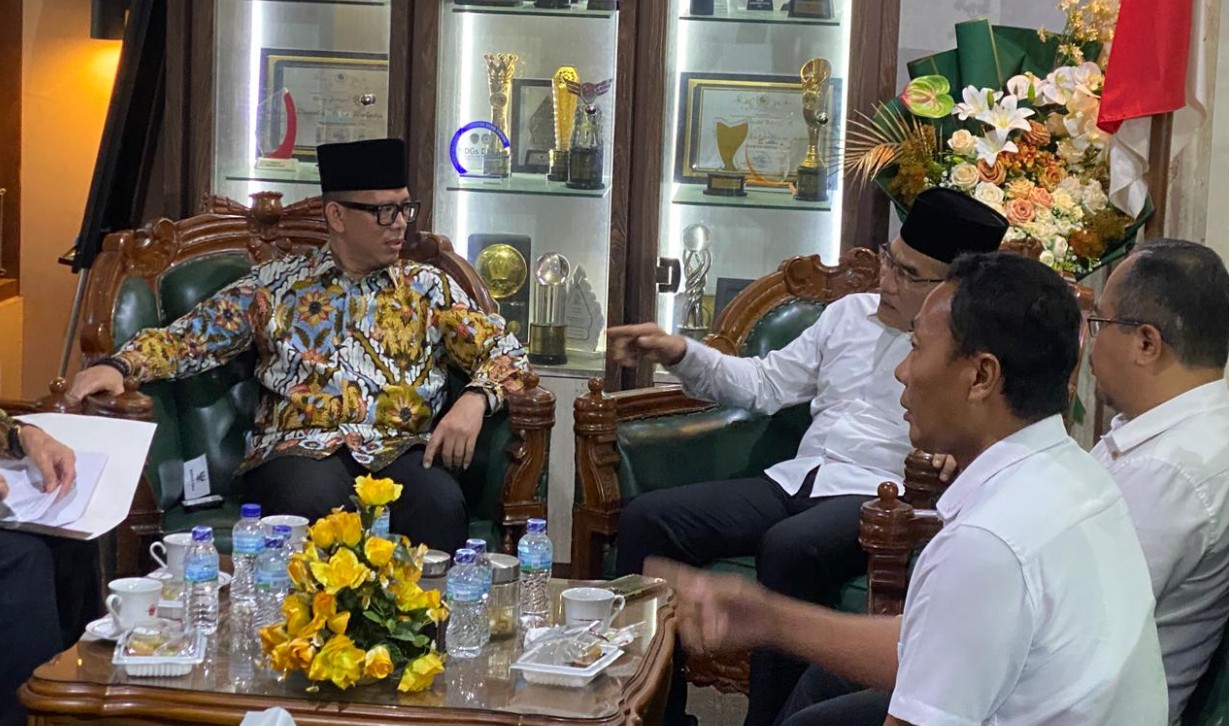 Bupati OKU berbaju batik dan Bupati Bantul berbaju putih duduk berdiskusi di ruang pertemuan dengan latar belakang lemari piala.