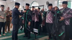 Bupati OKU, Teddy Meilwansyah (kiri), memasangkan jubah hakim hitam dengan lis hijau kepada seorang pria berbatik. Anggota Dewan Hakim lain mengenakan jubah mereka di latar belakang. Tanda 'DEWAN HAKIM' di lantai. Lokasi Islamic Center.
