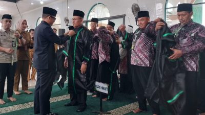 Bupati OKU, Teddy Meilwansyah (kiri), memasangkan jubah hakim hitam dengan lis hijau kepada seorang pria berbatik. Anggota Dewan Hakim lain mengenakan jubah mereka di latar belakang. Tanda 'DEWAN HAKIM' di lantai. Lokasi Islamic Center.