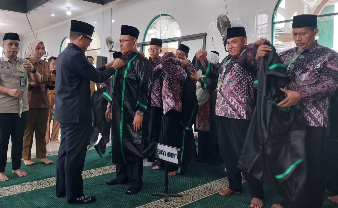 Bupati OKU, Teddy Meilwansyah (kiri), memasangkan jubah hakim hitam dengan lis hijau kepada seorang pria berbatik. Anggota Dewan Hakim lain mengenakan jubah mereka di latar belakang. Tanda 'DEWAN HAKIM' di lantai. Lokasi Islamic Center.