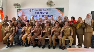 Foto grup besar guru-guru SMPN 19 OKU seragam PNS cokelat duduk dan berdiri di depan spanduk supervisi. Dua pria utama duduk di tengah depan. Beberapa guru membuat isyarat tangan OK.