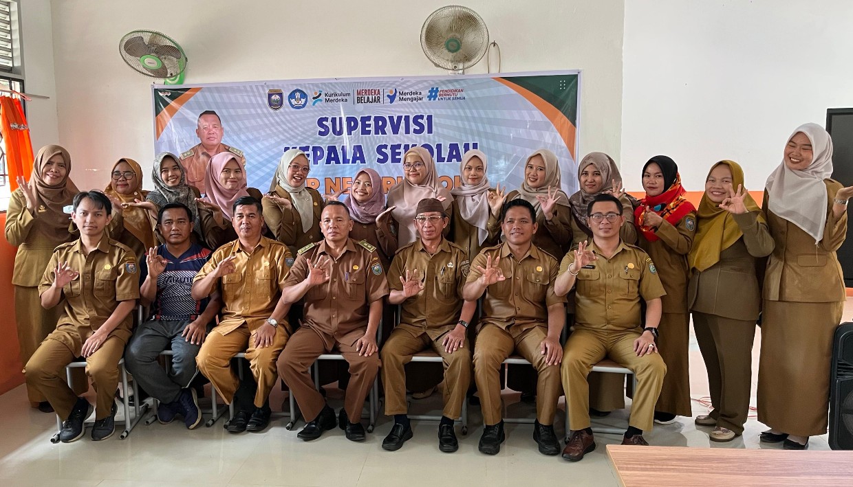 Foto grup besar guru-guru SMPN 19 OKU seragam PNS cokelat duduk dan berdiri di depan spanduk supervisi. Dua pria utama duduk di tengah depan. Beberapa guru membuat isyarat tangan OK.