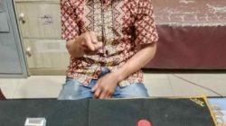 Seorang pria paruh baya mengenakan kemeja batik dan celana jins, duduk di depan meja hitam dengan wajah disensor. Di atas meja terdapat 17 paket plastik kecil berisi kristal putih (sabu), kotak rokok biru-putih, wadah merah, dan ponsel. Latar belakang kantor polisi.