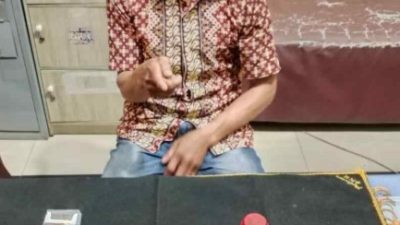 Seorang pria paruh baya mengenakan kemeja batik dan celana jins, duduk di depan meja hitam dengan wajah disensor. Di atas meja terdapat 17 paket plastik kecil berisi kristal putih (sabu), kotak rokok biru-putih, wadah merah, dan ponsel. Latar belakang kantor polisi.