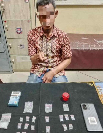 Seorang pria paruh baya mengenakan kemeja batik dan celana jins, duduk di depan meja hitam dengan wajah disensor. Di atas meja terdapat 17 paket plastik kecil berisi kristal putih (sabu), kotak rokok biru-putih, wadah merah, dan ponsel. Latar belakang kantor polisi.