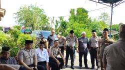 Sekelompok pria, sebagian besar berseragam Polisi RI dan satu berseragam Satpol PP, duduk dan berdiri bersama di area luar ruangan yang teduh di depan sebuah rumah. Seorang pria berbaju koko abu-abu dan peci hitam duduk di tengah, tampaknya sedang bercakap-cakap. Ada papan bunga dukacita di latar belakang.